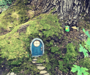 A tree root with a small blue fairy house door and stone path. A green toy animal with a tiny paper umbrella in place to the side of the door.