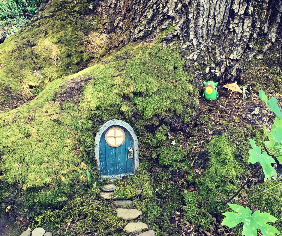 A tree root with a small blue fairy house door and stone path. A green toy animal with a tiny paper umbrella in place to the side of the door.
