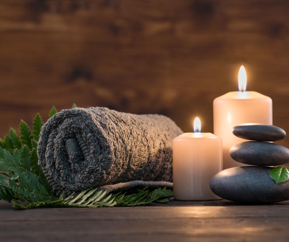 A rolled plush towel, two candles and three black stone on a dark wooden table.
