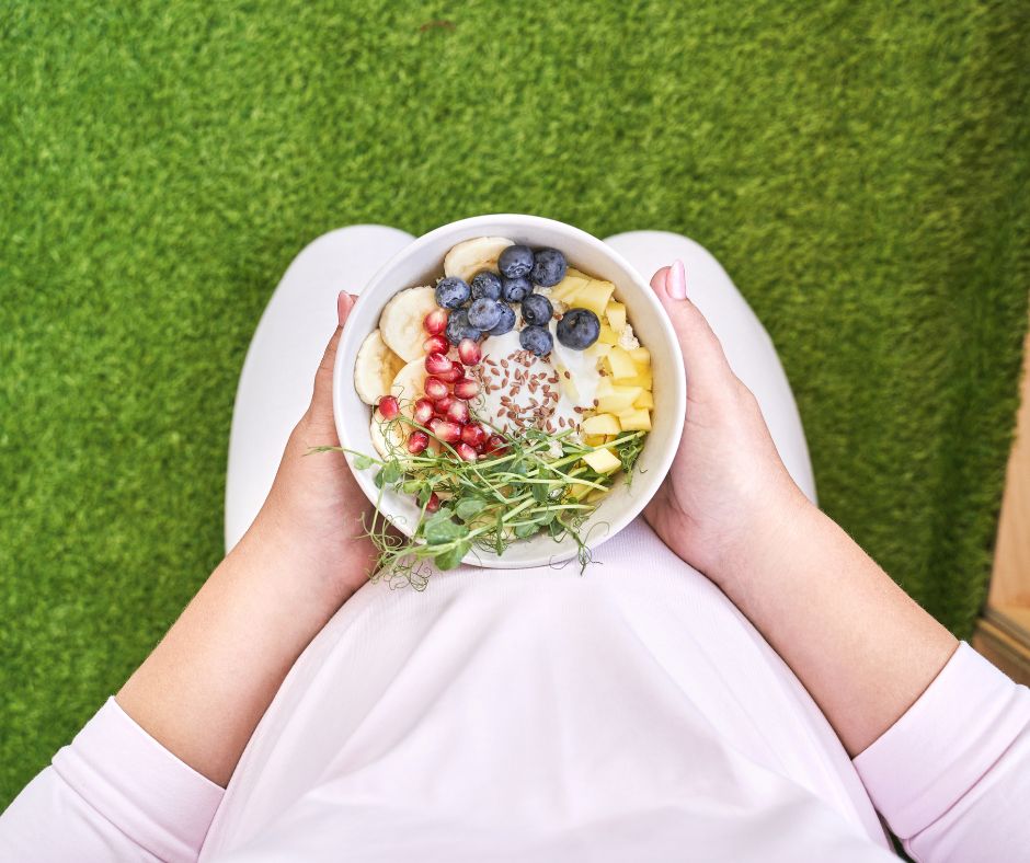 A person holding a bowl of sliced bananas, blue berries, pineapple, pomegranate seeds, pea microgreens, and yogurt topped with flax seeds.