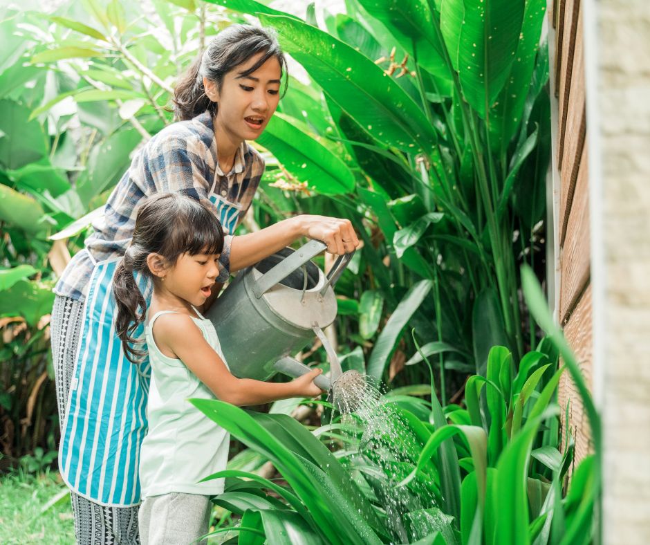 A woman and a girl using a watering can to water their large tropical outdoor plants.