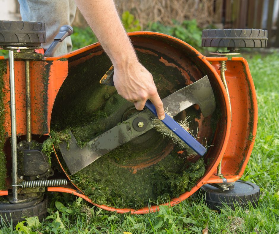 A hand using a scraper to clean the blade of an orange lawn mower as part of lawn mower winterization.