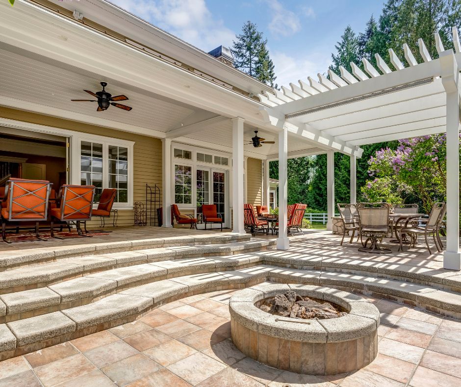 A white wooden freestanding pergola being used to extend the shady area of a patio for a dinning set next to a stone fire pit.