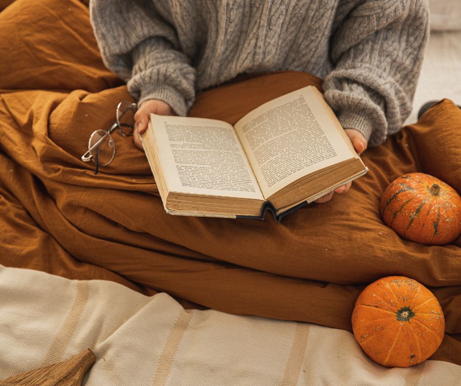 Cozy burnt orange blanket wrapped around a person wearing a grey cable knit sweater reading a book.