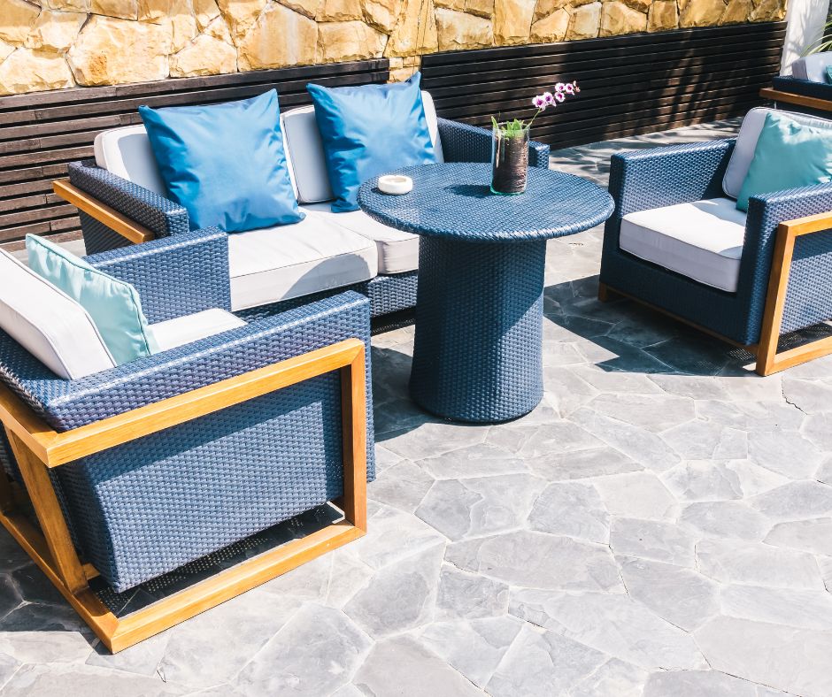 An outdoor seating area using a coastal style color palette of shades of light blue, wood, and seafoam white. Two chairs, a couch and round table with an orchid on a stone patio.