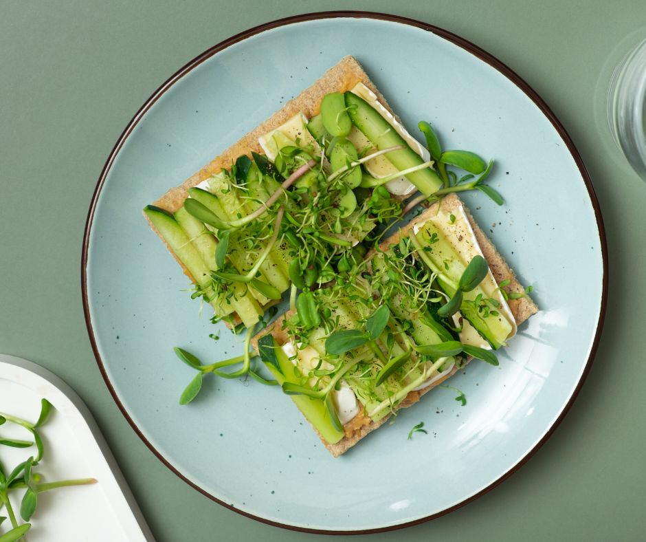 A light blue plate with two crackers on it. The crackers have slices of cheese, cucumber, and an assortment of green microgreens on them.