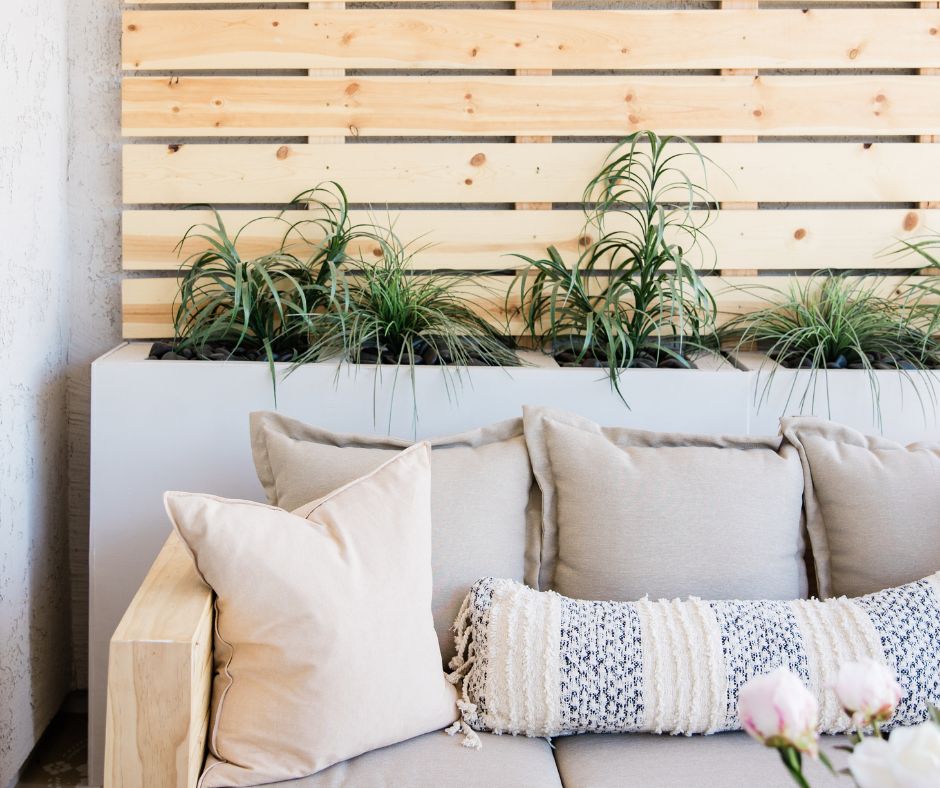An outdoor seating area featuring a neutral tone couch and pillows, a slated wood back drop and a planter filled with wispy green plants.