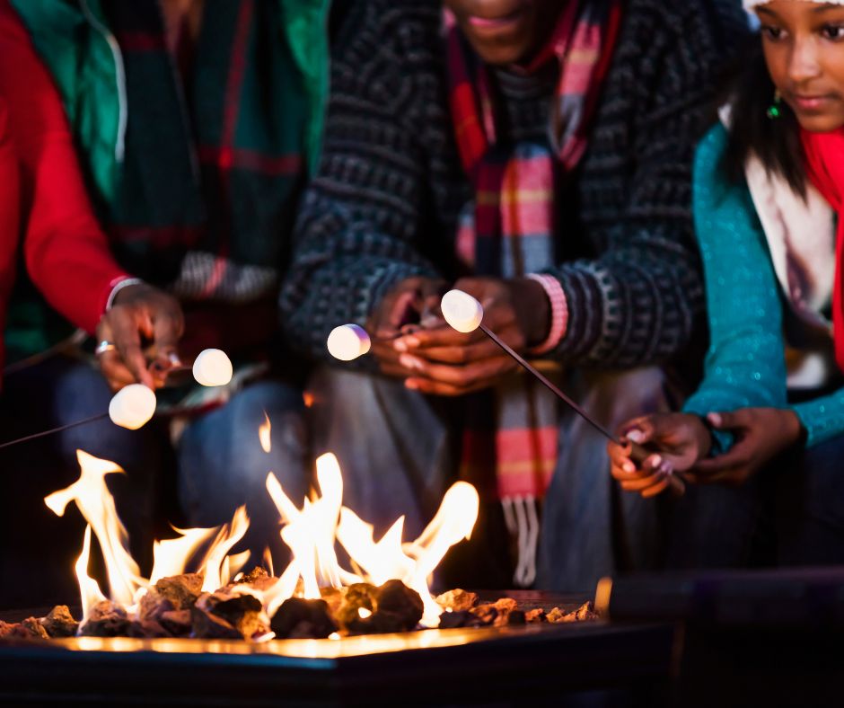 Family wearing colorful puffy coats, scarves and knit hats roasting marshmallows around a fire pit.