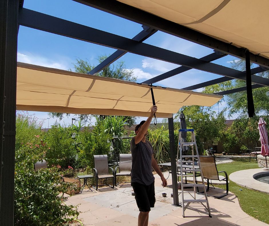 Man pulling beige redactable shade cloth close on a metal freestanding pergola installed to cover a stone patio.