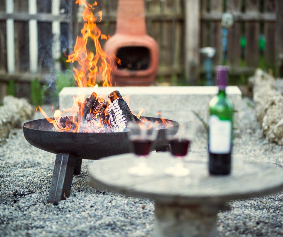 A metal fire pit with a fire burning in it. Behind it a clay chimenea. In the forefront a bottle of red wine with glasses.