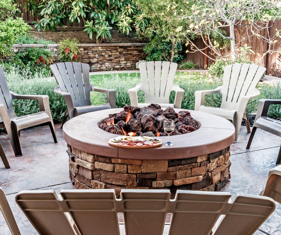 A brick fire pit with a pizza and a glass of red wine surrounded by beige Adirondack chairs.