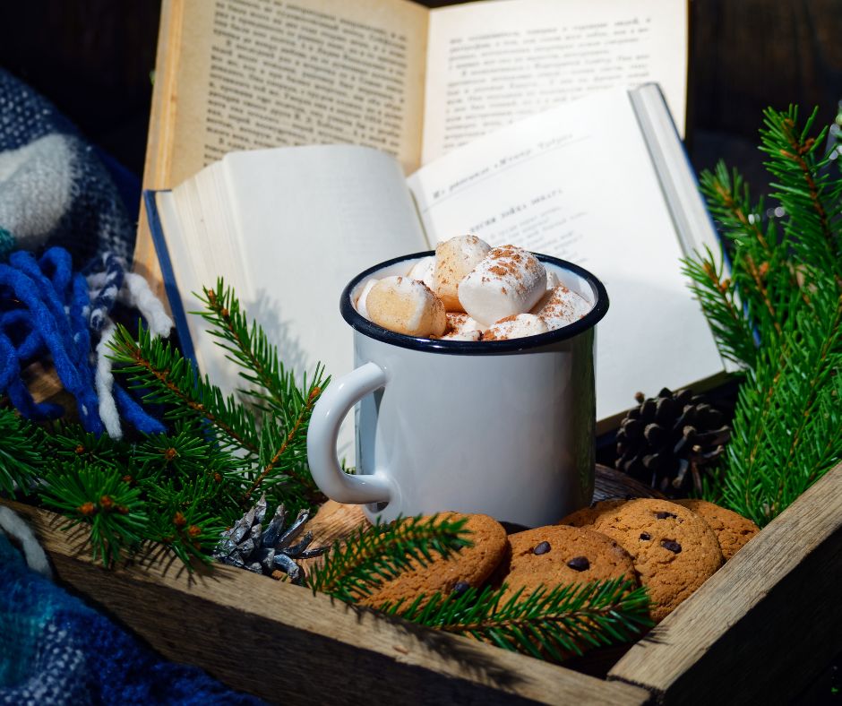 Hot cocoa with marshmallows in a white bug on a wood tray with cookies, books and evergreen branches.