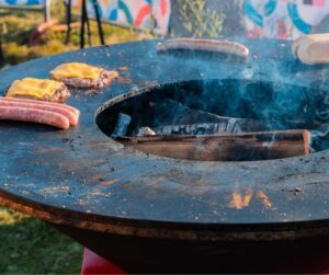A fire pit with a griddle attachment on which sausages and hamburgers with cheese are cooking.