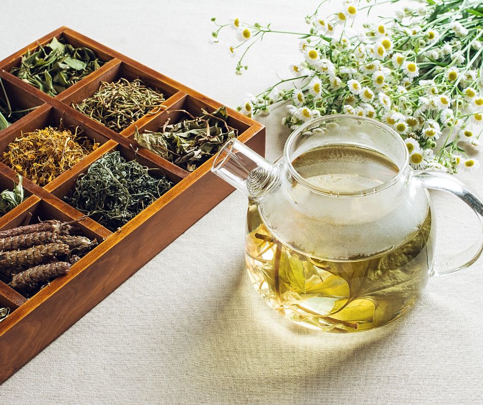 A glass tea pot next to fresh chamomile and and an assortment of dried herbal teas in a wooden box.