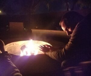 A man warming his hands at a the flames of a fire pit at night.