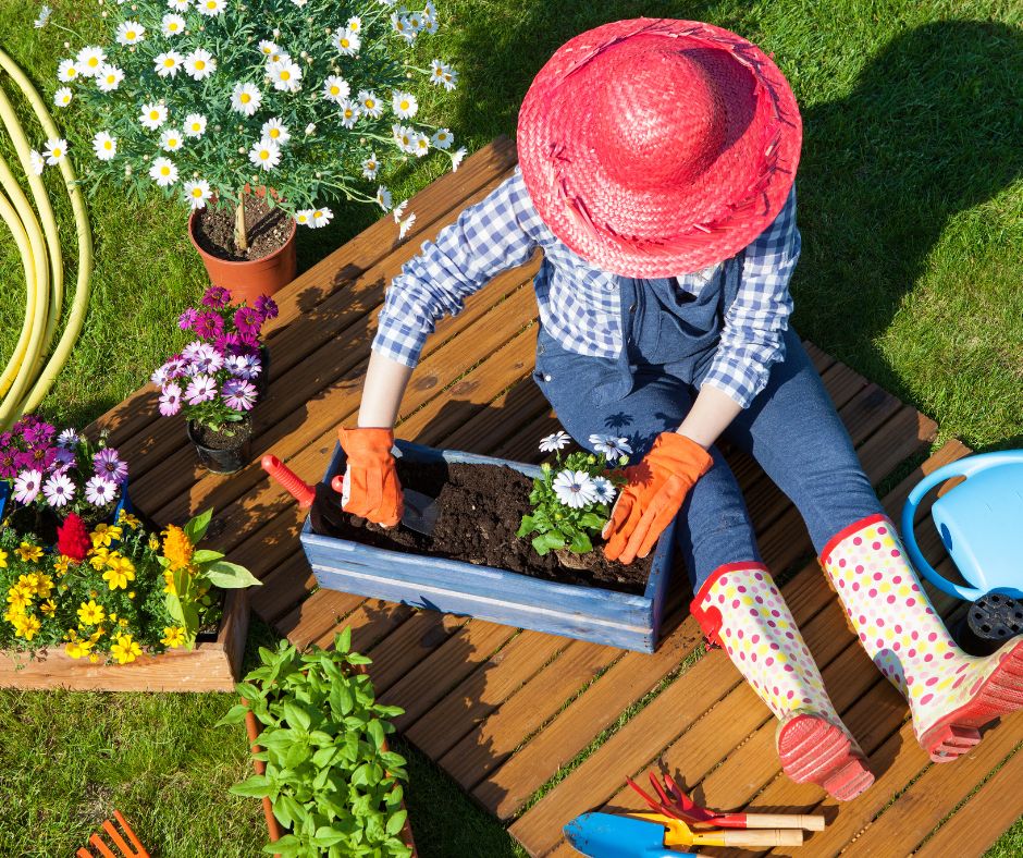 A gardener wearing a pink sun hat sitting on a wood deck potting up flowers.