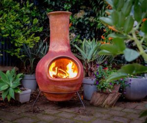 A clay chimenea on a brick patio surround by potted plants with a fire burning inside.