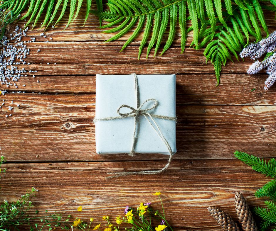 A present in a square box wrapped in white paper secured with natural brown twine. On a wooden table fringed with plant leaves.