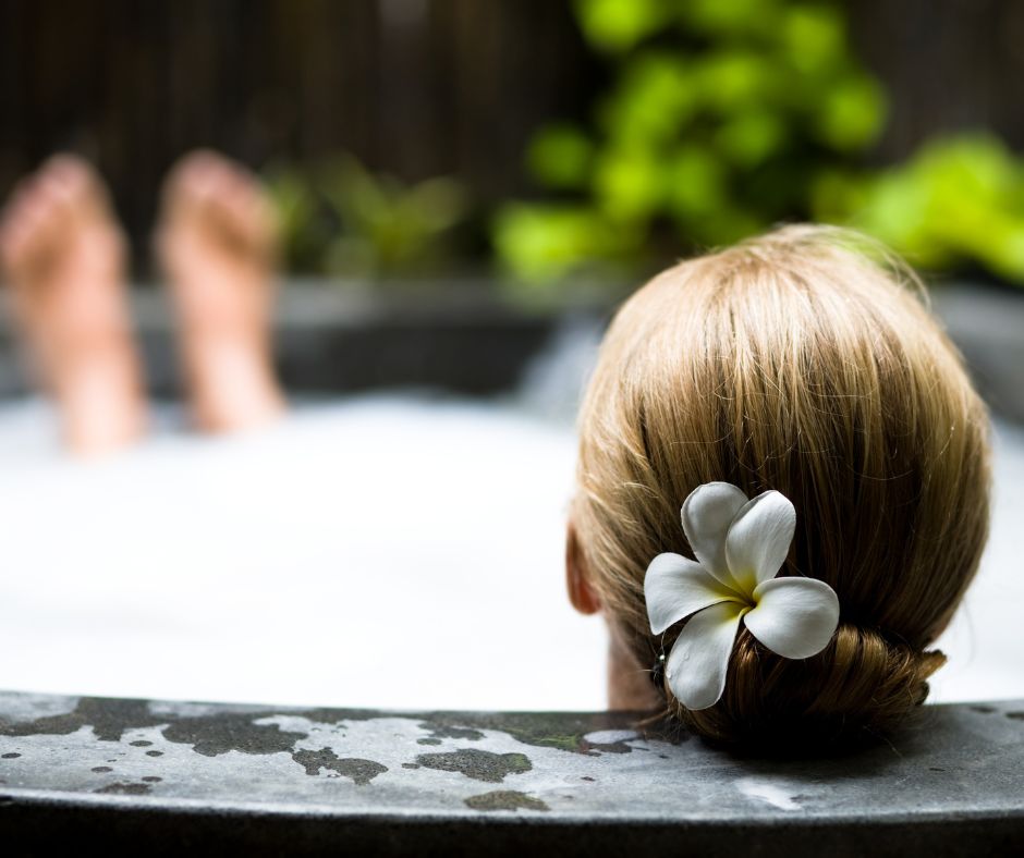 A person with a white flower in their hair soaking in a private hot tub surrounded by greenery.