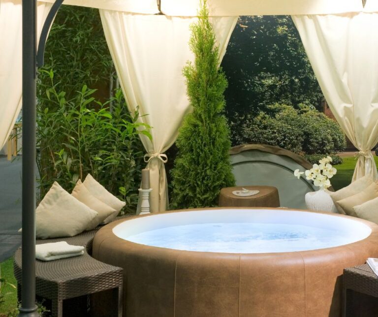 A private hot tub surrounded by large shrubs, a seating area and outdoor curtains.