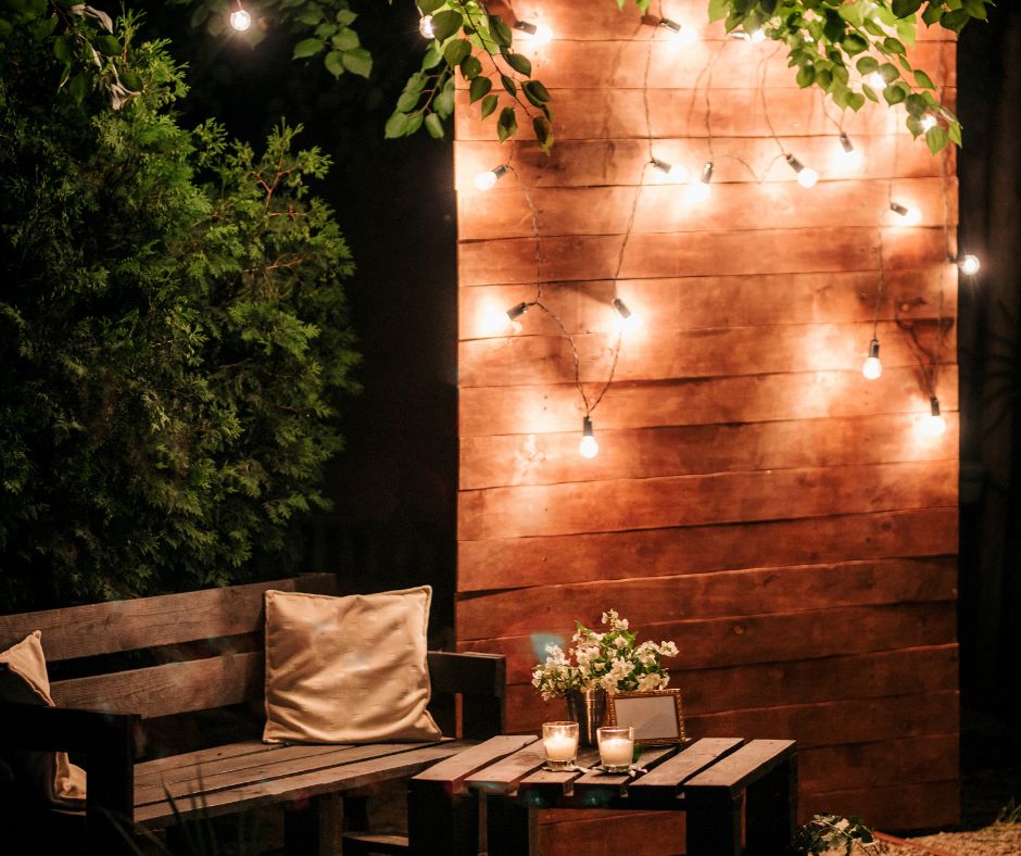 Warm string lights draped on wood panel next to a wood bench and table with two candles.