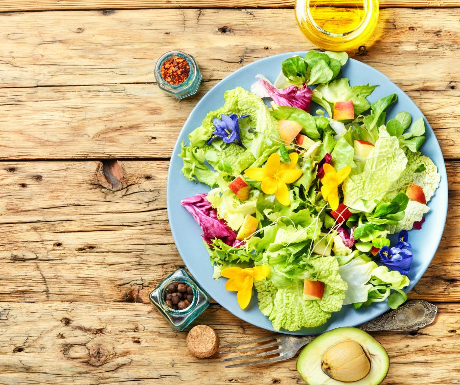 A colorful salad topped with edible flowers on a wood table surrounded by spices, oil, avocado, and a fork.