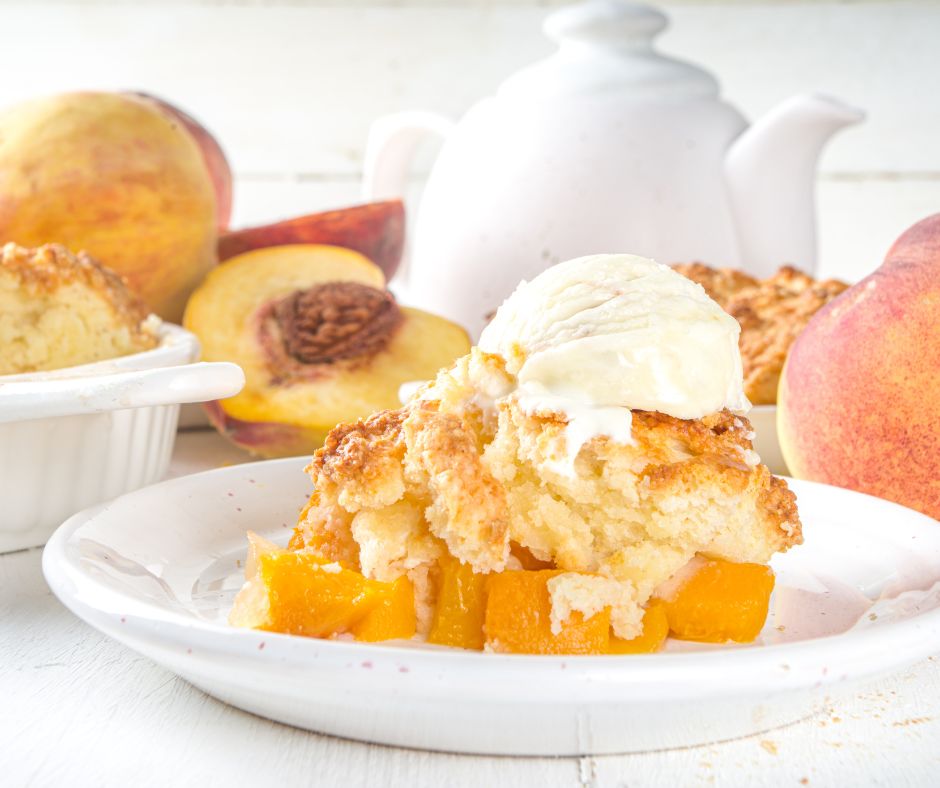Peach cobbler with ice cream on a white plate surrounded by fresh peaches on a white table.