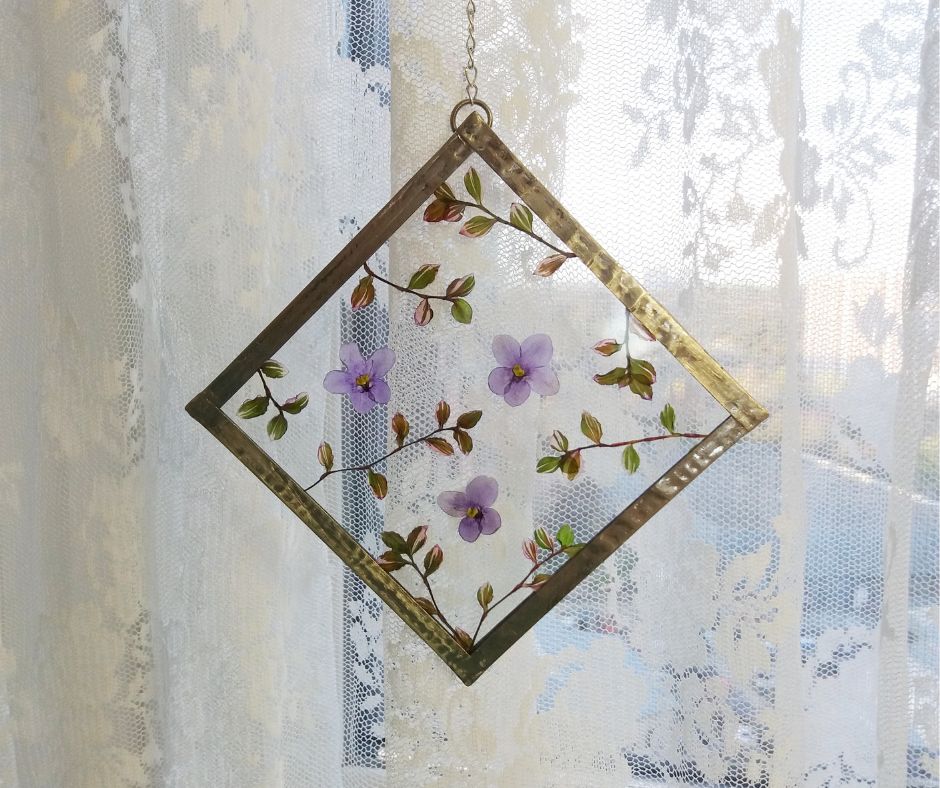 A diamond shaped pressed flower light catcher in front of white lace curtains. 