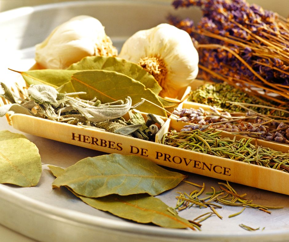 A wood tray with the words "Herbes De Provence" on the side in black. Compartments of the tray have different dried herbs in them including, sage, rosemary, bay leaf, thyme and garlic bulbs.