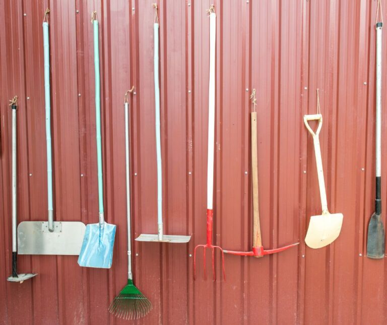 A selection of long handled garden tools hanging on a wall of a red shed.
