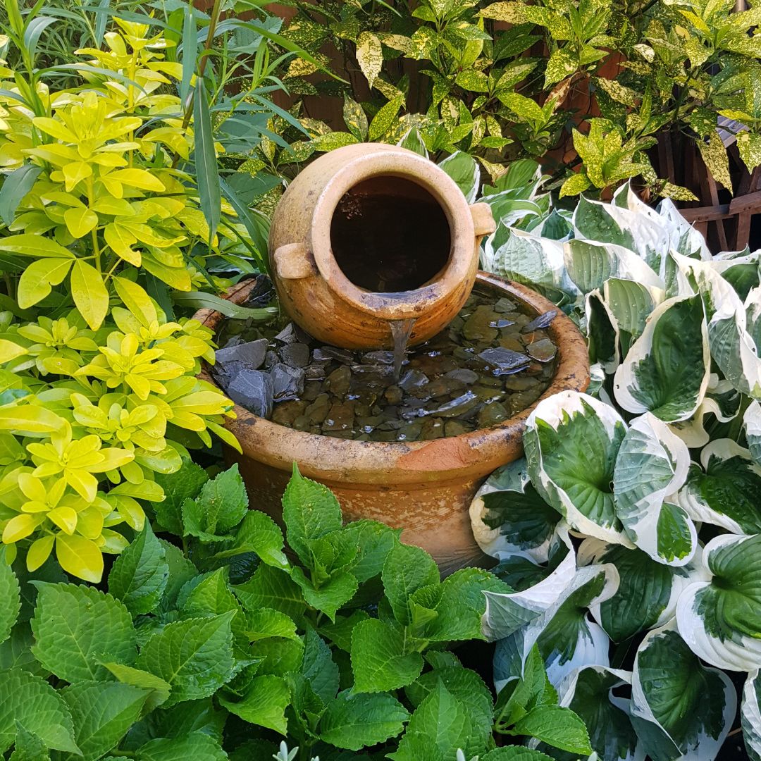 A terracotta pot water fountain surround by lush garden greenery.