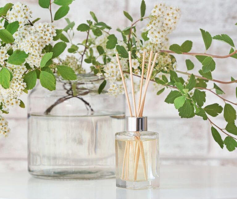 A glass essential oil reed diffuser on a white table with a vase of blooming foliage. In front of a white brick wall.