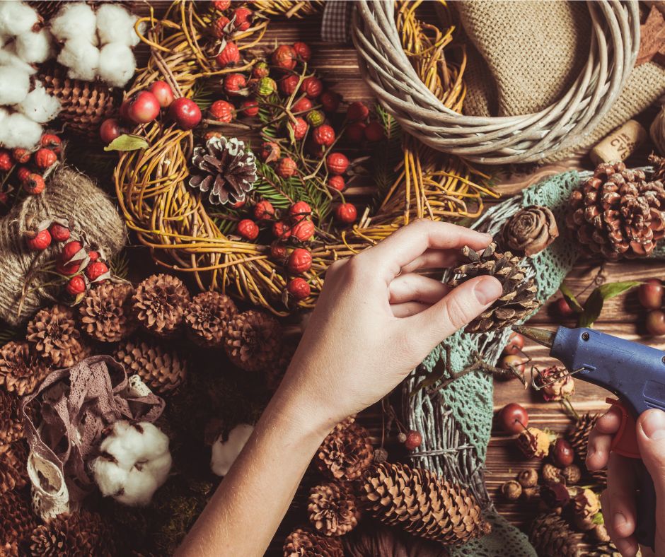 A hand using a glue gun to attach decorations onto a variety of dried grapevine wreaths. A classic natural decoration.