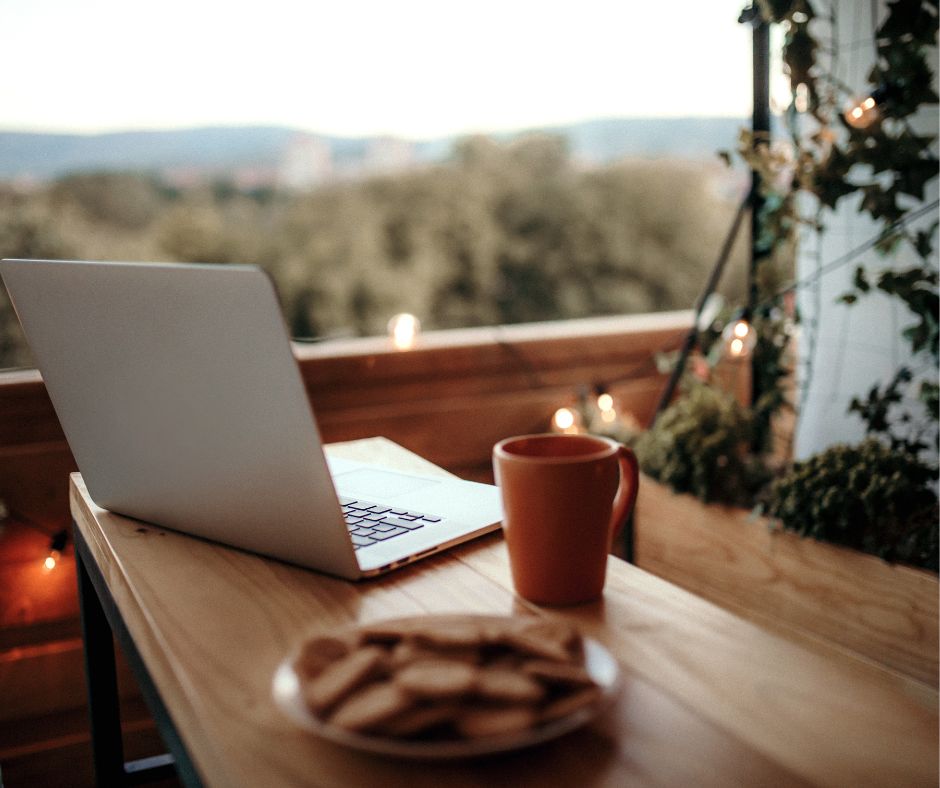 A laptop on a wood table with a mug and cookies in a hybrid living space.