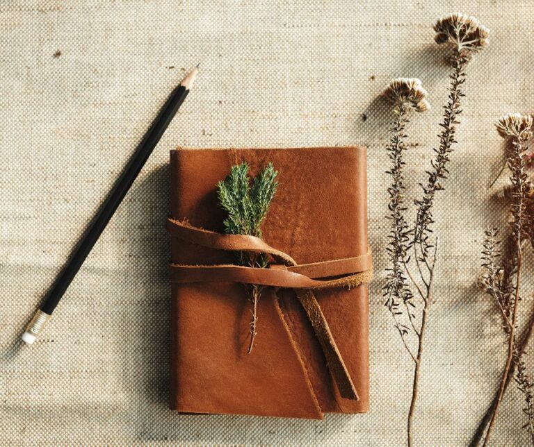 A small brown notebook with dried plants and pencil on a white linen table cloth.