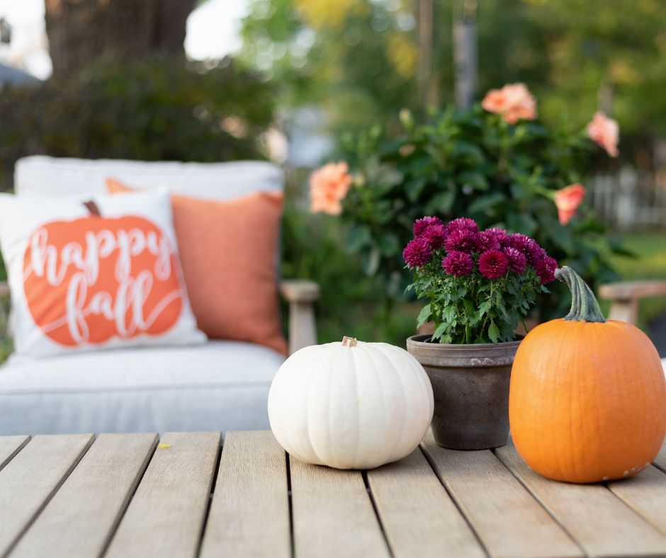 A patio decorated for fall. Wood table with small pumpkins and a pot with red mum flowers. A chair with a festive happy fall pillow.