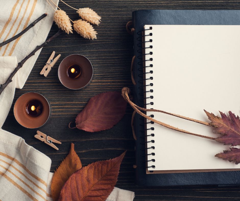 A spiral notebook on top of a black bound journal on a wooden table with leaves and candles.