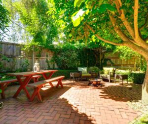 A brick patio with a red bench and table sitting area surrounded by greenery and a privacy fence.