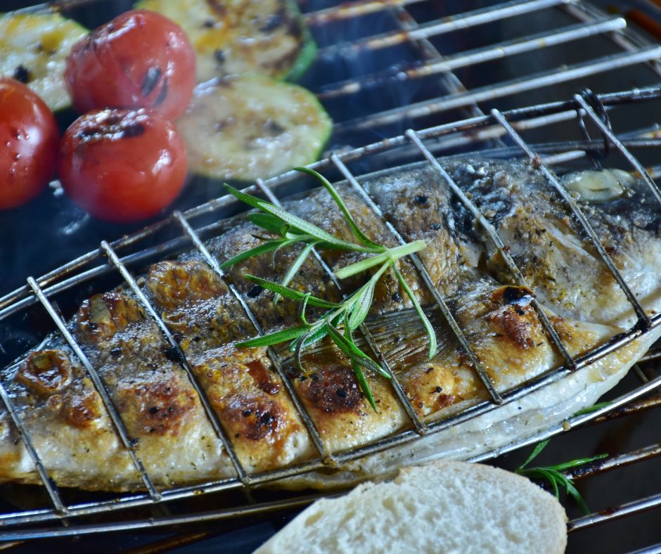 Fish in a grill basket one a grill next to tomatoes.