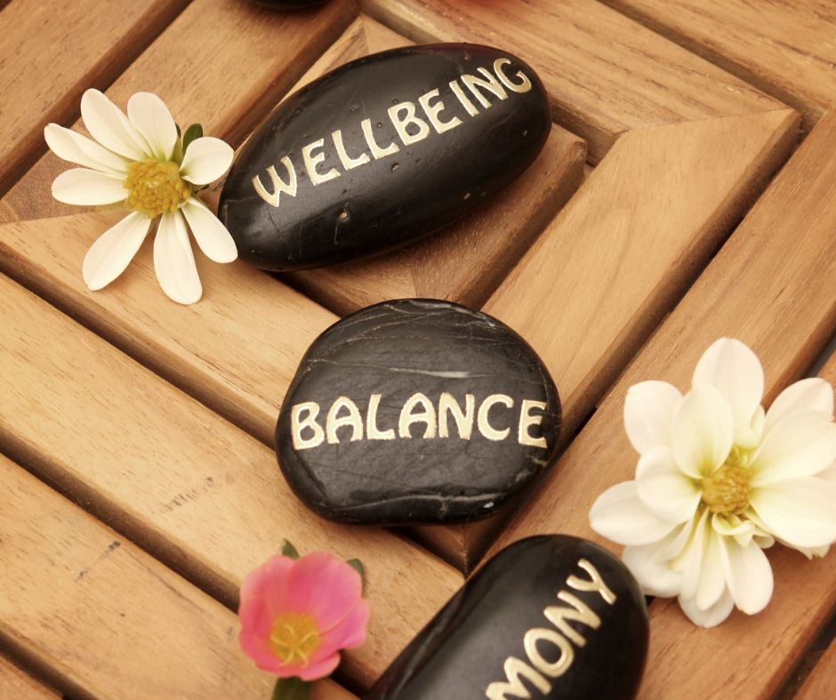 The words "wellbeing, ballance and harmony" written in white on black stones next to flowers on a wooden table.