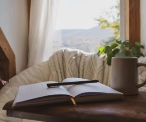 A cozy journaling nook. An open journal on a wooden table with coffee next to an open window.