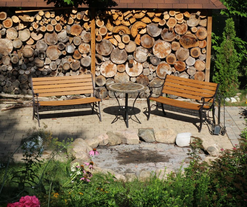 A fire pit siting area with two benches sheltered by a large stack of cut wood.