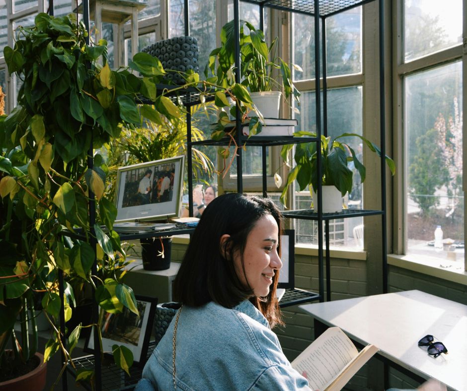 A woman reading a desk surround by houseplants in front a window.