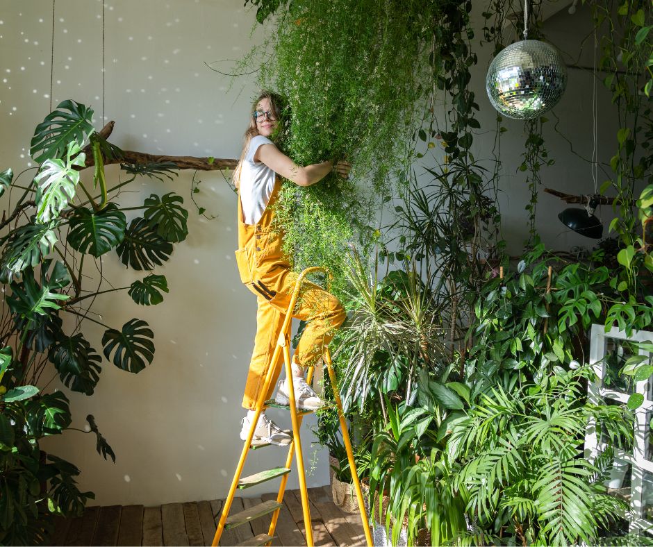 A woman in yellow overall hugging a large hanging plant in a sunroom.