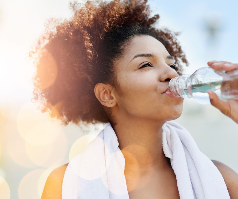 A woman drinking water from a bottle with a white towel around her neck.