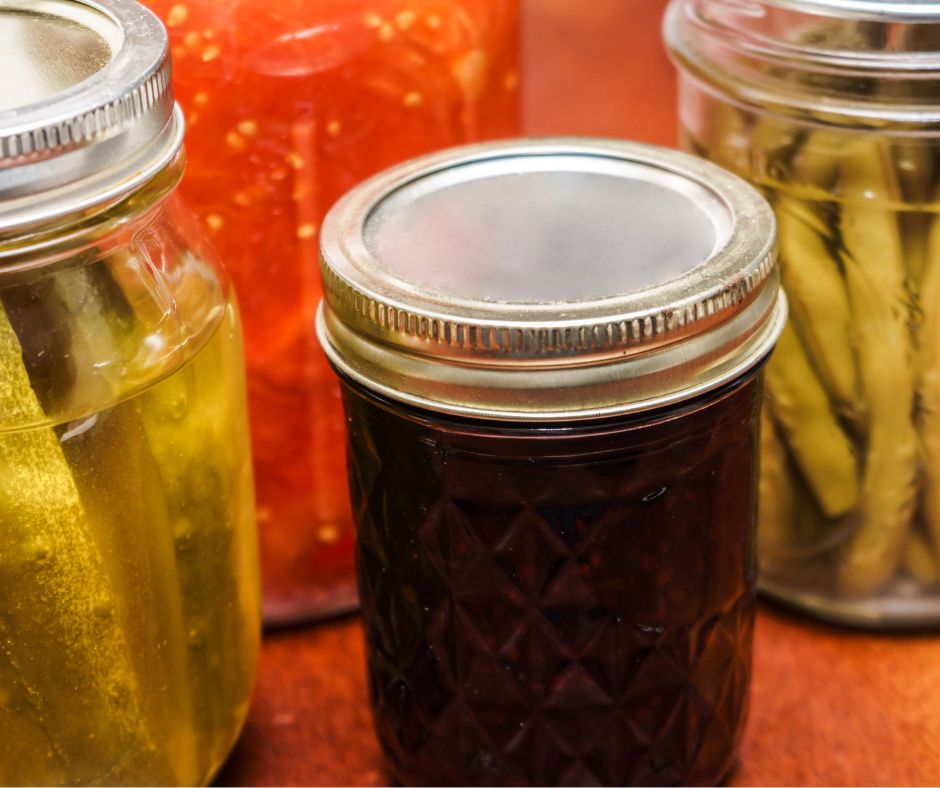 Jars of dark purple jelly, green beans, pickles, and tomatoes.
