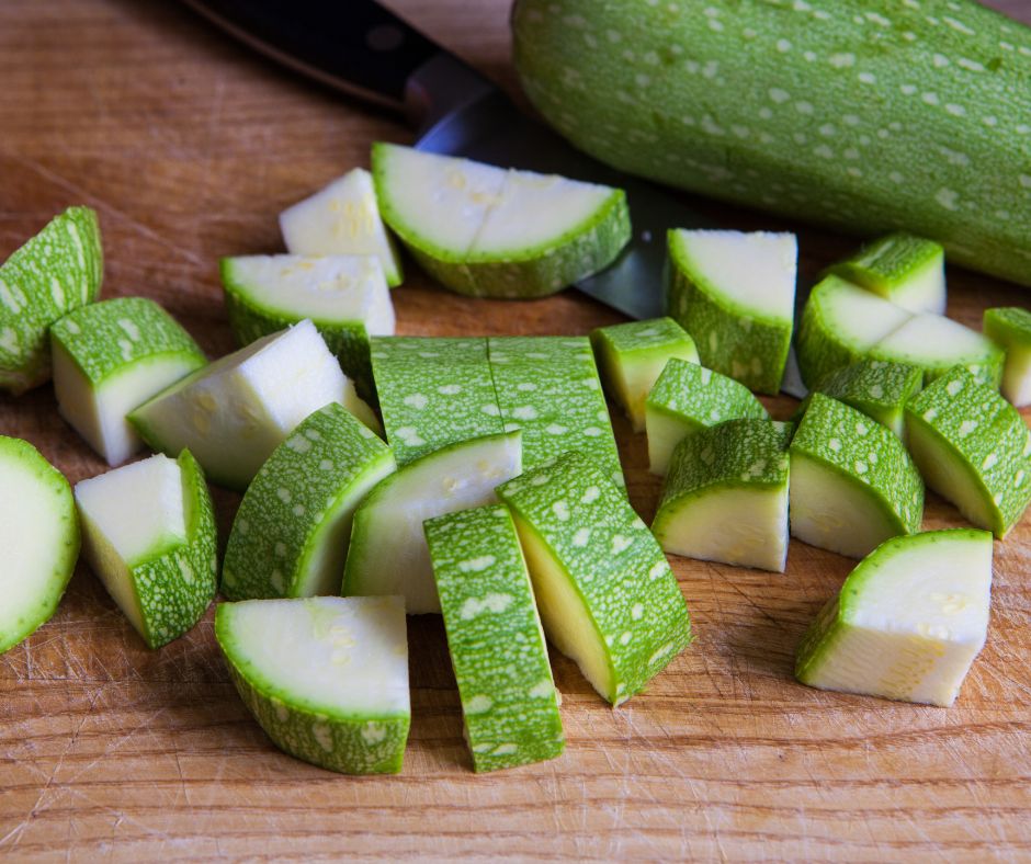 Cut pieces of zucchini on a cutting board in front of a whole squash harvested from a garden.
