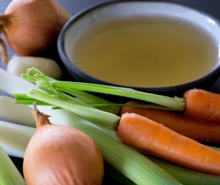 Onions, leeks and carrots next to a bowl of broth.