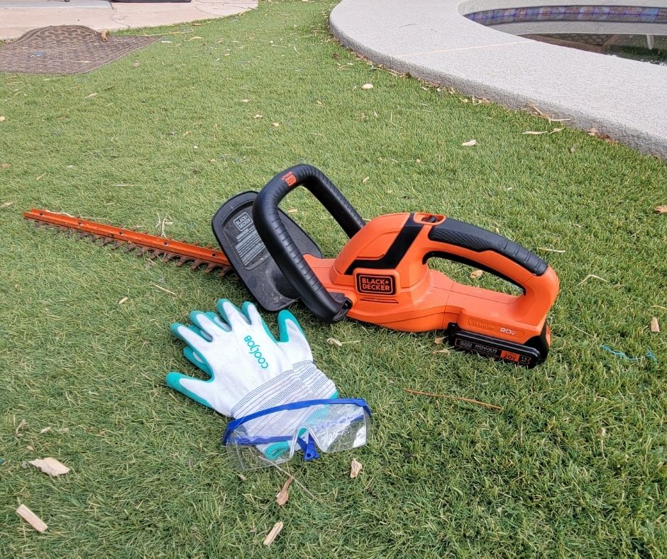 White and green garden gloves next to a cordless trimmer on green turf.