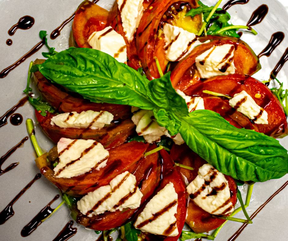 A Capri salad of red sliced tomatoes, mozzarella cheese, herbs, avocado, fresh green basil and drizzled with rich brown balsamic vinegar.